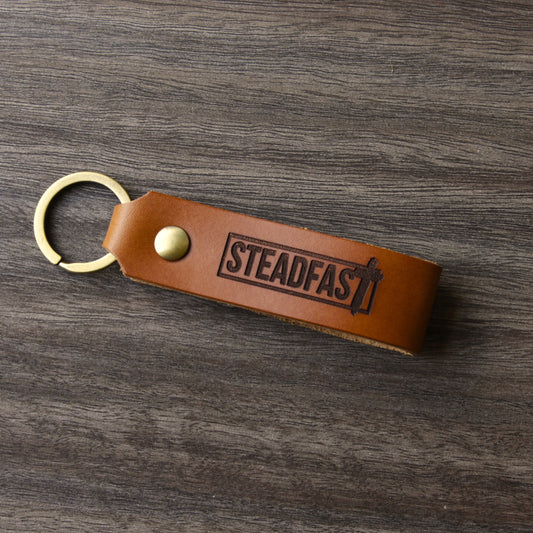 A brown leather keychain with the word 'Steadfast' printed on it, placed on a wooden surface.